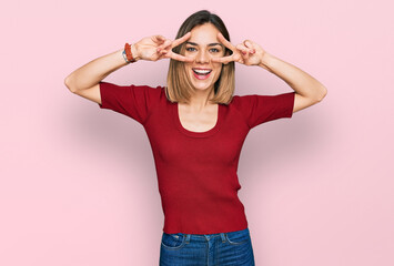 Young blonde girl wearing casual clothes doing peace symbol with fingers over face, smiling cheerful showing victory