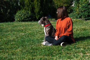 Una linda y alegre mujer caucásica está sentada en el parque, junto a su perro marrón de la raza American Stafford Terrier