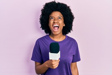 Young african american woman holding reporter microphone angry and mad screaming frustrated and...