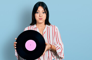 Young hispanic girl holding vinyl disc puffing cheeks with funny face. mouth inflated with air, catching air.