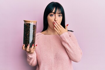 Young hispanic woman holding jar with coffee beans covering mouth with hand, shocked and afraid for...