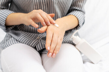 Close-up of an unrecognizable woman applying natural moisturizer cream to her hands while sitting on the bed at home. Moisturizing, nourishing, regeneration, care and protection dry hand skin.