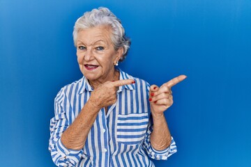 Senior woman with grey hair standing over blue background smiling and looking at the camera pointing with two hands and fingers to the side.