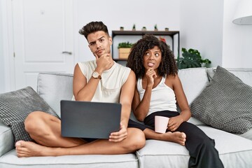 Young interracial couple using laptop at home sitting on the sofa thinking worried about a question, concerned and nervous with hand on chin