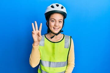 Beautiful brunette little girl wearing bike helmet and reflective vest showing and pointing up with fingers number three while smiling confident and happy.