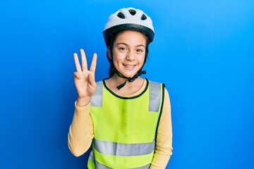 Beautiful brunette little girl wearing bike helmet and reflective vest showing and pointing up with...