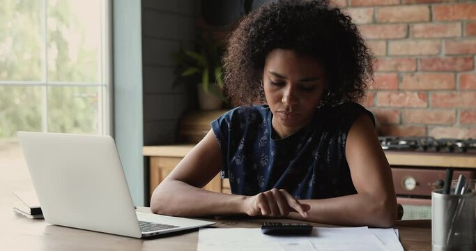 Busy young African woman pay utility bills in online internet bank app using laptop check taxes sum on calculator plan domestic budget economy. Thoughtful female do financial paperwork at home office