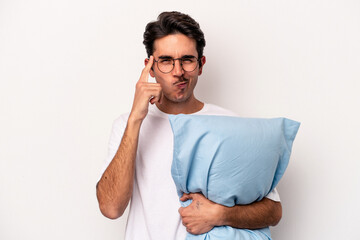 Young caucasian man wearing pajamas holding a pillow isolated on white background pointing temple...