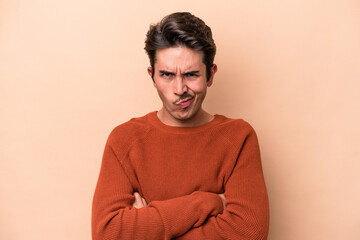 Young caucasian man isolated on beige background frowning face in displeasure, keeps arms folded.