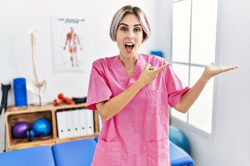 Young nurse woman working at pain recovery clinic amazed and smiling to the camera while presenting with hand and pointing with finger.