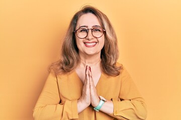 Middle age caucasian woman wearing casual clothes and glasses praying with hands together asking for forgiveness smiling confident.