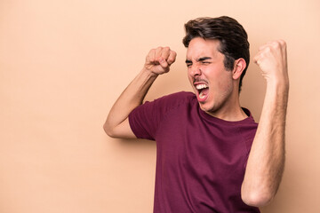 Young caucasian man isolated on beige background raising fist after a victory, winner concept.