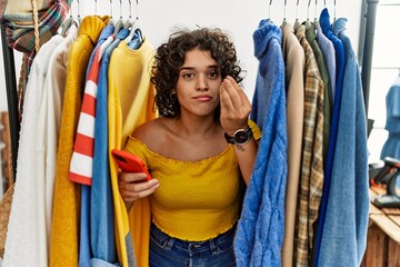 Young hispanic woman searching clothes on clothing rack using smartphone doing italian gesture with hand and fingers confident expression