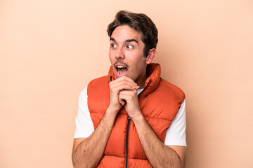 Young caucasian man isolated on beige background praying for luck, amazed and opening mouth looking to front.