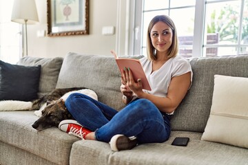Obraz premium Young caucasian girl smiling happy sitting on the sofa with dog reading book at home.