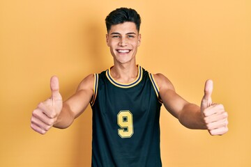 Young hispanic man wearing basketball uniform approving doing positive gesture with hand, thumbs up...