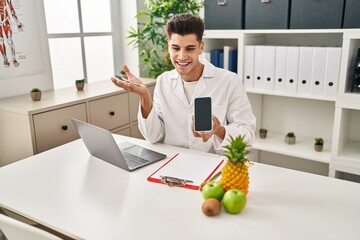 Young hispanic man wearing dietician uniform showing screen smartphone at clinic