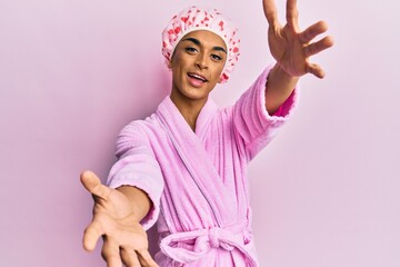 Hispanic man wearing make up wearing shower towel cap and bathrobe looking at the camera smiling with open arms for hug. cheerful expression embracing happiness.