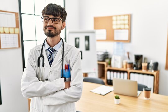 Hispanic man with beard wearing doctor uniform and stethoscope at the office smiling looking to the side and staring away thinking.