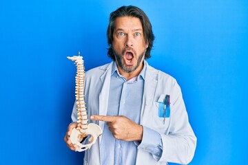 Fototapeta premium Middle age handsome traumatologist man holding anatomical model of spinal column afraid and shocked with surprise and amazed expression, fear and excited face.