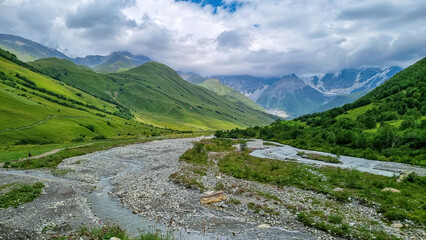 Fototapeta na wymiar Patara Enguri River flowing down the a valley with view on the Shkhara Glacier in the Greater Caucasus Mountain Range in Georgia, Svaneti Region, Ushguli. Snow-capped mountains in the back. Wilderness