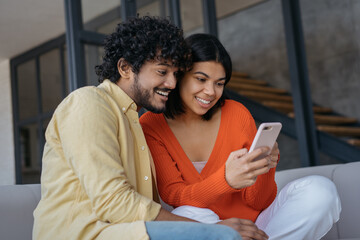 Smiling multiracial friends using mobile phone shopping online relaxing at home. Happy Indian man...