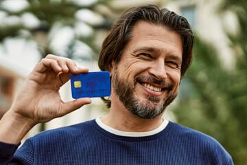 Middle age handsome man holding credit card outdoors at the park