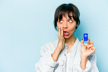 Young hispanic woman holding a batterie isolated on blue background is saying a secret hot braking news and looking aside