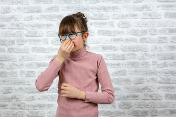 Portrait of frustrated school girl pinching nose with disgust on his face due to bad smell isolated over gray brick wall background. smelling something stinky and disgusting, intolerable smell.