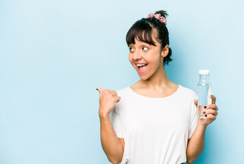 Young hispanic woman holding a bottle of water isolated on blue background points with thumb finger away, laughing and carefree.