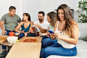 Group of young hispanic friends eating italian pizza sitting on the sofa. Woman smiling happy and using smartphone at home.