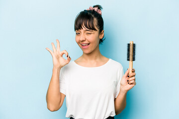 Young hispanic woman holding a brush hair isolated on blue background cheerful and confident showing ok gesture.