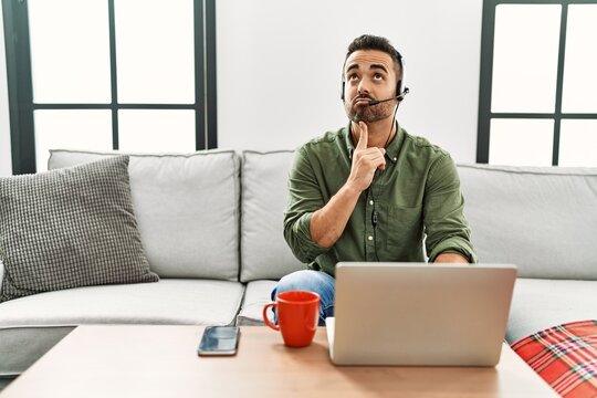 Young hispanic man with beard wearing call center agent headset working from home thinking concentrated about doubt with finger on chin and looking up wondering