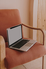 Blank screen laptop on comfortable peachy lounge chair. Home interior design. Copy space mockup....