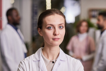 Young contemporary female physician or general practitioner in lab coat looking at camera while standing in hospital against colleagues