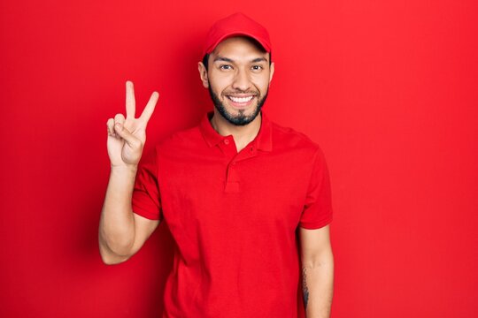 Hispanic man with beard wearing delivery uniform and cap smiling looking to the camera showing fingers doing victory sign. number two.
