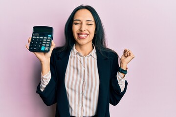 Young hispanic girl showing calculator device screaming proud, celebrating victory and success very excited with raised arm - obrazy, fototapety, plakaty