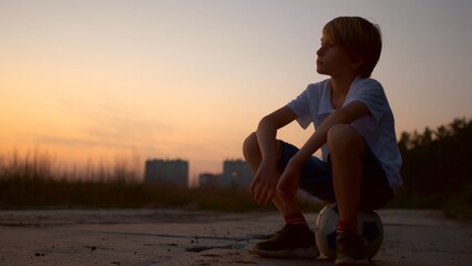 A teenage boy in the evening in the city sits on a ball and dreams of the future sports career of a football player, future soccer celebrity. The childhood of a great soccer player.