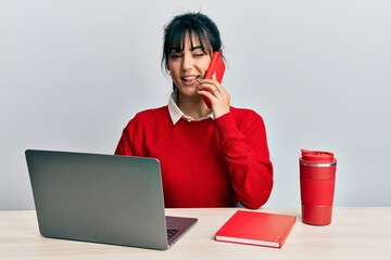 Young brunette woman with bangs working at the office talking on the smartphone winking looking at...