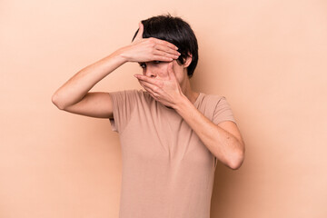 Young caucasian woman isolated on beige background blink at the camera through fingers, embarrassed covering face.