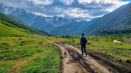 Fototapeta premium A man walking with a dog toward the Shkhara Glacier in the Greater Caucasus Mountain Range in Georgia, Svaneti Region, Ushguli. Stray dog. Wanderlust, hiking. Humans best friend. Outdoor activities.