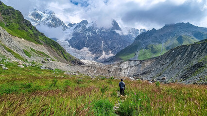 Fototapeta na wymiar A man hiking and enjoying the panoramic view on the Shkhara Glacier in the Greater Caucasus Mountain Range in Georgia, Svaneti Region, Ushguli. Wanderlust, Alpine pasture. Blooming flower.