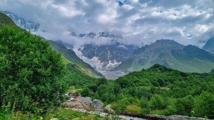 Fototapeta premium Patara Enguri River flowing down the a valley in the Greater Caucasus Mountain Range in Georgia, Svaneti Region, Ushguli. The Shkhara Mountain is covered by clouds. Snow-capped mountains.Glacier