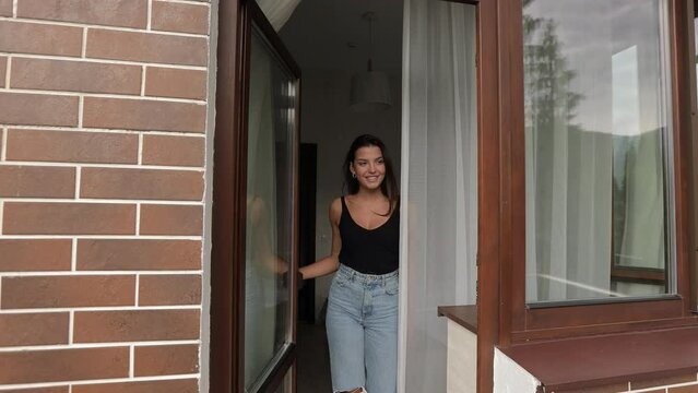 Beautiful smiling woman in casual wear opening door on balcony from her comfy hotel room. Portrait of happy female tourist. Travelling and people concept.