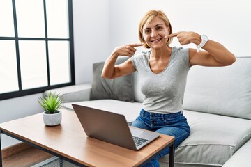 Middle age blonde woman using laptop at home smiling cheerful showing and pointing with fingers teeth and mouth. dental health concept.