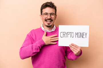 Young caucasian man holding a crypto money placard isolated on beige background laughs out loudly keeping hand on chest.