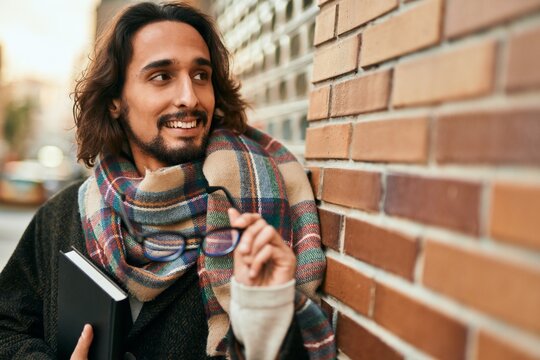 Young hispanic man smiling happy holding glasses and book at the city.