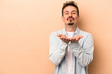 Young caucasian man isolated on beige background folding lips and holding palms to send air kiss.