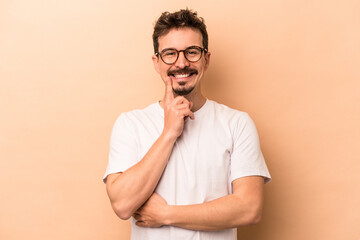 Young caucasian man isolated on beige background smiling happy and confident, touching chin with hand. - obrazy, fototapety, plakaty