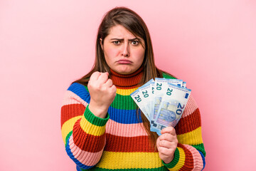 Young caucasian overweight woman holding banknotes isolated on pink background showing fist to camera, aggressive facial expression.
